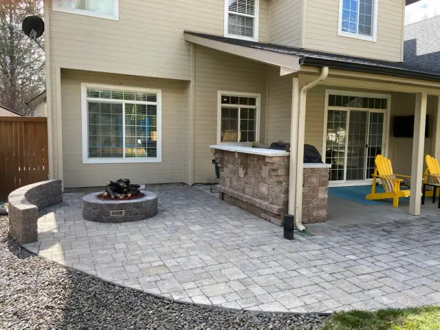 A completed backyard patio with stone pavers, featuring a built-in stone bar and seating area, a round fire pit, and yellow Adirondack chairs. The patio is attached to a beige house with large windows, providing a comfortable and inviting outdoor living space.